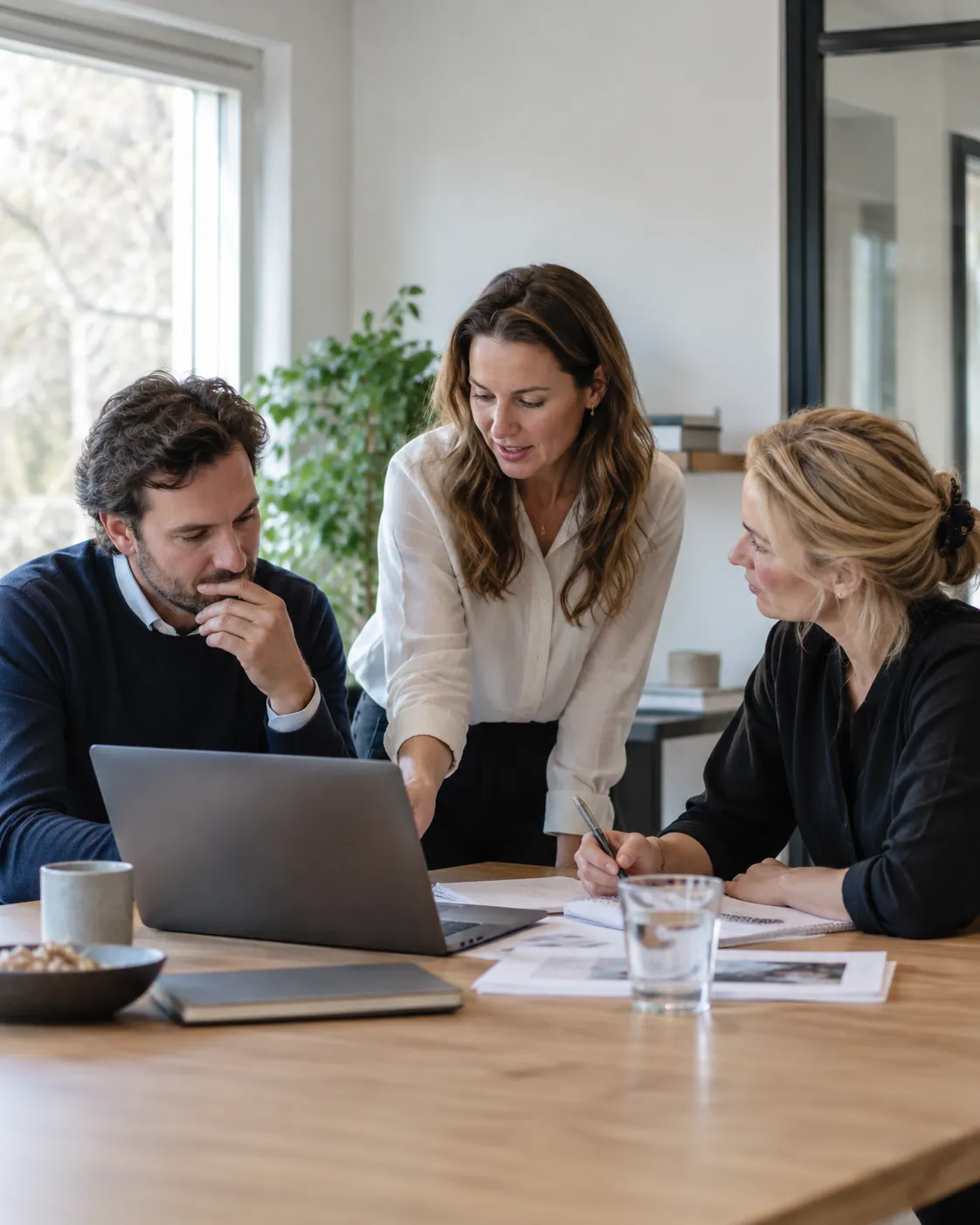 Three colleagues discussing documents around a laptop in a bright office.